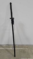 Full katana sword with black wrapped handle, metal guard, and black scabbard standing upright.
