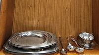 Stack of three oval metal serving platters and several serving utensils including ladles and gravy boat arranged in front.