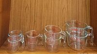 Photo of 7 Pyrex glass measuring cups arranged on wood background with visible handles and spouts.