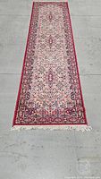 Full view of the traditional red and cream runner rug laid out flat on a grey concrete floor, showing its full length and fringed ends.