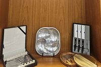 Overall view of shelf showing the butter spreaders box, pewter serving bowl, Cutco knives, and wooden cheese cutting board set with knives.