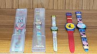 Wide view of all 5 Swatch watches including 2 still in packaging and 3 loose with colorful designs and straps.