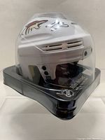 White mini hockey helmet with Arizona Coyotes logos, autographed on top and side by Anthony DeAngelo, Christian Dvorak, Lawson Crouse, and Ryan MacInnis. Displayed on black plastic stand with clear protective visor.