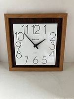 Full frontal view of the Bulova square wall clock with teak wood trim, white face, and black numerals and hands.