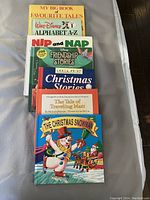 Photo showing a stack of seven children's hardcover books with titles visible on covers including 'My Big Book of Favourite Tales,' 'Walt Disney Alphabet A-Z,' 'Nip and Nap Disney Friendship Stories,' 'Large Print Christmas Stories,' 'The Tale of Traveling Matt,' and 'The Christmas Snowman.'