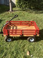 Full side view of the children's wagon sitting on grass showing wooden base, red wooden sides, and metal handle.