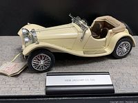 Side view of the 1/24 scale cream-colored 1938 Jaguar SS100 Roadster model showing brown interior, chrome grille, and wire-spoke wheels on the black display base with plaque.