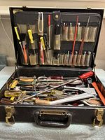 Open black tool case displaying assorted hand tools including screwdrivers with yellow and red handles, pliers, and files in top and bottom compartments.