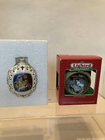 Two Christmas religious glass ornaments displayed with their original boxes: on left a white oval Mary and Joseph ornament, on right a red box holding a round lighted ornament.