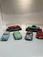 Six small model cars arranged with two in clear plastic display cases with plaques, others positioned in front.