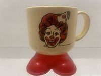 Front side view of off-white plastic cup featuring Ronald McDonald's illustrated face with party hat. Large red clown feet form the base of the cup.
