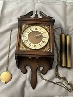 Front view of full clock face, ornate metal corner decoration, included pendulum on left and two brass weights with chains on right.