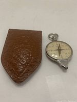 Vintage metal compass alongside brown textured leather carrying case on white background.