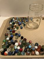 Tray with assortment of vintage marbles of varying sizes and colors, next to empty glass jar.