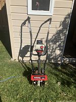 Front view of Yard Machines 31cc cultivator showing handles, engine, and rotary tiller blades on grass against house siding.