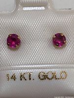 Close-up photo of the ruby stud earrings mounted on 14K gold settings in a jewelry holder labeled '14 KT. GOLD'
