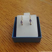 Front view of the 14K gold earrings with pear-shaped tanzanite stones displayed on a white holder inside a small blue box on a wooden surface.