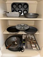 Shelving with baking pans and party serving utensils arranged on three shelves: muffin pan, rectangular pan, round pans, divided serving pan, springform pan, scissors and pie servers on metal tray.