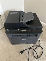 Full view of Brother all-in-one printer model DCP-L25500W with power cable extended on carpeted floor.