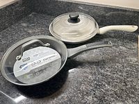 Image showing two ceramic non-stick fry pans on countertop: one IKO with gray handle, black ceramic interior, and a BKLYN Steel Co with white handle and glass lid.