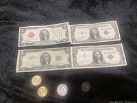 Four US paper bills including $1 and $2 denominations, some with red and blue seals, laid out on black fabric with four coins positioned below bills.