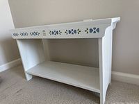 Front angled view of white wooden bench with bottom storage shelf and stenciled floral designs on side rails.
