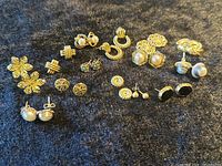 Full overview of 14 paired vintage women's earrings on a dark fabric background, showing various styles and decorative gold-tone metal details including faux pearls, rhinestones, and textured finishes.