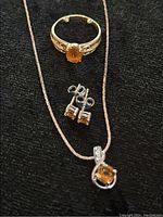 Photo shows gold-tone ring with orange citrine gemstone, silver-tone necklace with citrine pendant and clear stones, and pair of citrine stud earrings on black background.