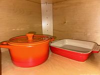 Side view of the orange Parini Dutch oven and red square Le Creuset baking dish inside a wooden cabinet.