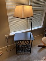 Side table with black metal frame and beige fabric lamp shade turned on, placed near window