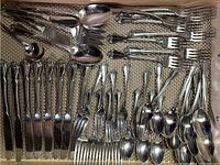Top view of arranged flatware piled in drawer, showing knives, forks, spoons, spreaders, and serving forks and spoons in stainless steel with ornate handles.