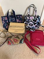 Overview of five purses and bags on a flat surface, showing different styles and colors including floral, tan, red quilted, and multicolor patterns.
