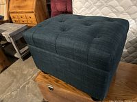 Front and top view of blue-green tufted storage ottoman placed on wooden chest, showing fabric texture and lid shape.