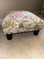 Angular side view showing floral upholstery, tufted top, and black round legs of the footstool.