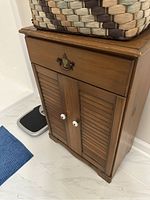 Front view of the vintage hamper cabinet showing the drawer with brass handle and double doors with white ceramic knobs.