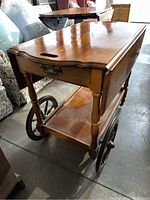 Angle view of vintage wooden tea cart showing polished surface, scalloped drop leaf with slot handle, turned legs, large wooden spoked wheels, and a lower shelf.