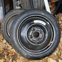 Photo showing two spare tires stacked outdoors on a bed of leaves. Tires mounted on black steel rims with visible lug nut holes.