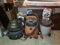 Photo showing three shop vacuums side by side with hoses and accessories on floor and countertop in background.