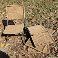 Four outdoor lawn chairs with woven seats and backrests arranged on grass. One chair is upright while others are stacked or leaning. Metal frame and tan woven material visible. Some wear and fraying noted.