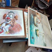 Photo showing two framed paintings side by side; one is a colorful clown portrait, the other is a seascape with boats.