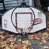 Front view of the Huffy Sports basketball backboard with hoop and net outdoors on ground with dry leaves around.
