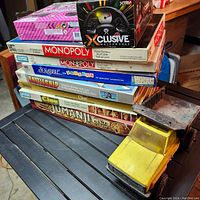 Stack of classic Parker Brothers boardgames (Monopoly Anniversary and standard editions, Scrabble for Juniors, Battleship, Clue, Jumanji) alongside toy vehicles (yellow truck and boxed Xclusive Motorworks car) and a pink boardgame box.