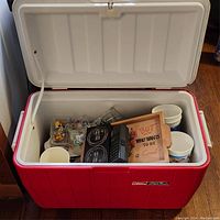 Red Coleman Polylite cooler opened to show contents including cups, glassware, and a small wooden sign.