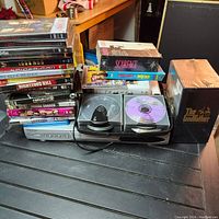 Dual-disc black DVD player placed in front with CDs and two VHS boxed sets labeled Scarface and The Godfather showing wear