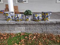 Front view of five ceramic flower pots, four empty and one with green artificial plant, all with yellow and blue floral motif.