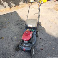 Overall view of the Honda gas lawn mower showing the red Honda engine cover, black mower deck, wheels, grass catcher, and broken handle.