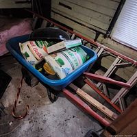 Side view of the blue Jackson wheelbarrow loaded with fertilizer bags and a Weber grill cover. Shows wheelbarrow plastic tub and orange metal frame.