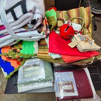 Photo showing the folded no-sew throw kit blanket, crocheted multi-color blanket, striped fabric curtains, red fabric, floral fabric, red metal jingle bell, and tied fabric coasters.