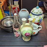 Lot of various vintage and modern decorative items including vases, bottles, ceramic jar, and metal bowl on a dark surface.