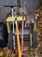 Overall shot of lot outdoors, showing all tools leaning against a stacked black plastic stand with yellow measurement stickers, on a leaf-covered surface near a building.